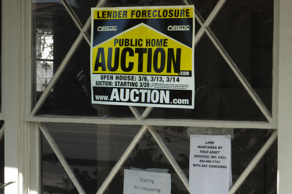 Window looking into a darkened foreclosed home. A yellow sign says "LENDER FORECLOSURE" and "PUBLIC HOME AUCTION." Smaller paper signs say "Warning No Trespassing" and note a lawn maintenance company.