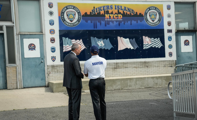 Mayor Eric Adams standing with his back to the camera, facing a poster reading "Rikers Island NYCD".