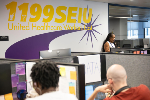 In the 1199 SEIU office, people work in at desks with a large 1199 SEIU: United Healthcare Workers East logo on the wall.