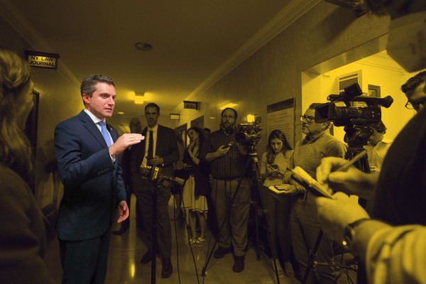 Senator James Skoufis is shown speaking to a group of reporters in a hallway.