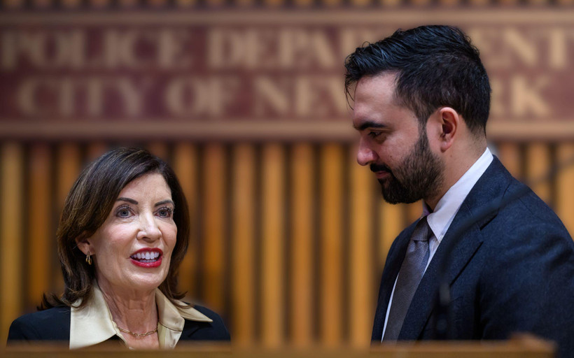 NYC Mayor Zohran Mamdani, in profile, looks at Governor Kathy Hochul, who looks outwards. The two stand close to each other behind a podium.