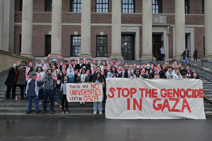 Photo of about 40 protestors on the steps of a Harvard building. All students are holding their hands up with their palms painted red. Two large banners read "THERE ARE NO UNIVERSITIES LEFT IN GAZA" and "STOP THE GENOCIDE IN GAZA."