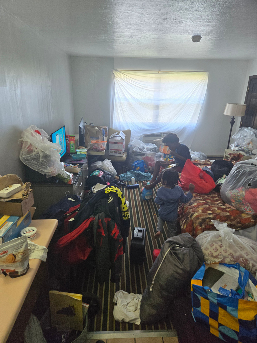 Taylor and Samir watch television in the motel room filled with suitcases and bags. A bedsheet acts as a curtain for the window.