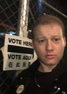 Will Bredderman stands in front of sign reading Vote Here / Vote Aquí and a chainlink fence