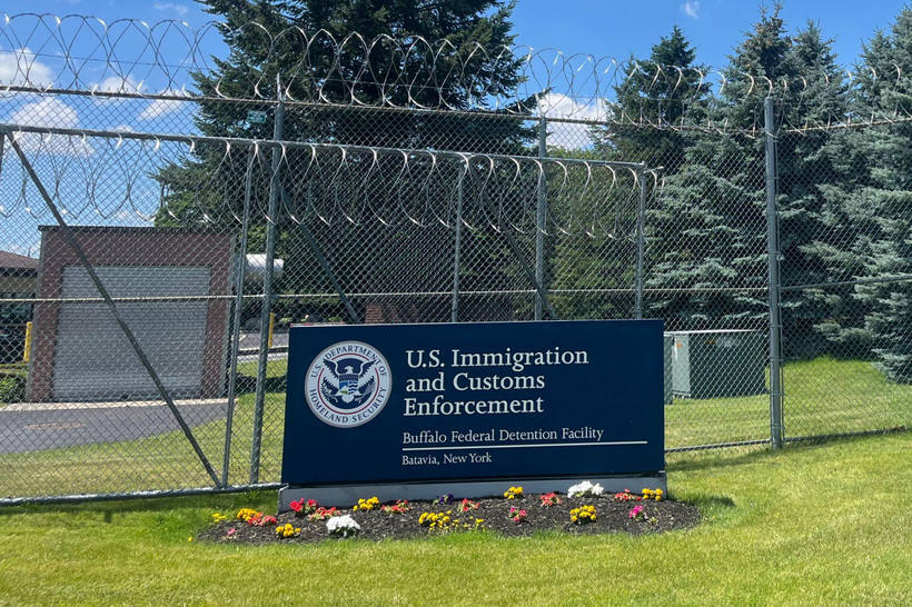 A blue sign reading "U.S. Immigration and Customs Enforcement, Buffalo Detention Center, Batavia New York" sits on the grass in front of a tall metal fence with barbed wire at the top.