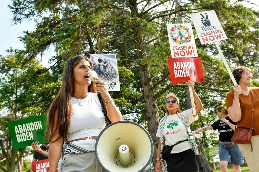 A woman protester shouts into a large megaphone in the foreground. A few older protesters stand behind her with signs reading "Gaza love," "Abandon Biden--ceasefire now" and have flower and peace sign imagery.