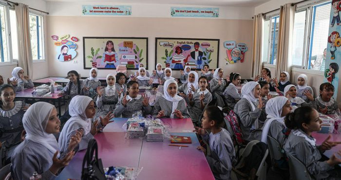 A classroom of two dozen girls chatter and gesture. Behind them are colorful posters of kids reading.