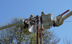 Two men in hard hats work on a utility pole from cherry picker buckets in Massena, New York.
