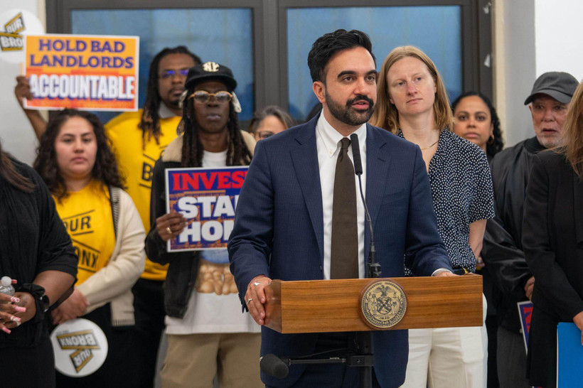 At a press conference, Mayor Zohran Mamdani stands speaking at a podium, behind and to his left stands Cea Weaver, director of the New York City Mayor's Office to Protect Tenants. Behind them stand people holding signs bearing the words "Hold bad landlords accountable" and "Invest in stables homes."