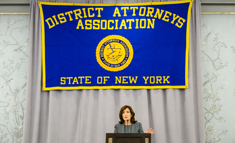 New York Governor Kathy Hochul speaks in front of a blue and yellow banner that reads  District Attorneys Association of the State of New York.