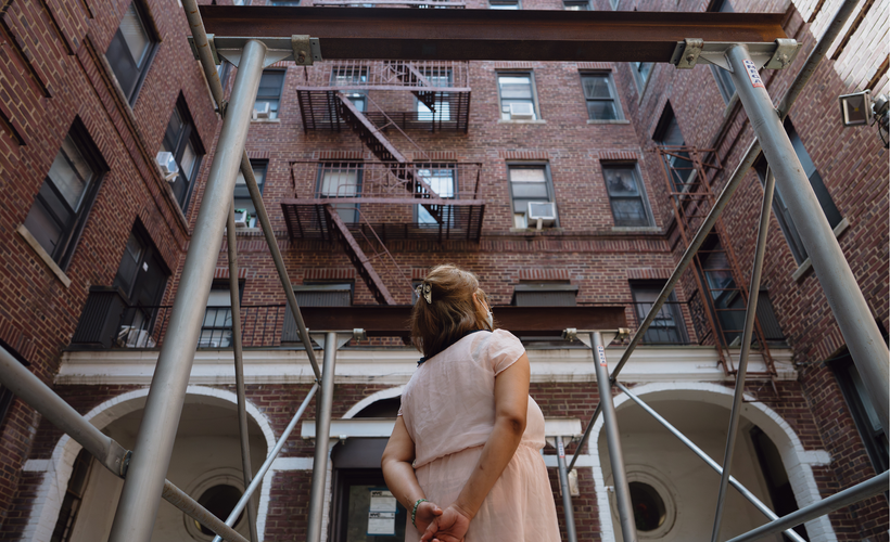 A woman stands outside a brick building, looking away from the camera, with her hands clasped behind her back.